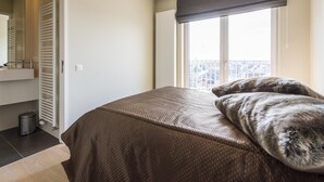 A bedroom with a bed, a window with a view, a radiator, and a door leading to a bathroom. - Beautiful apartment by the sea (Blankenberge)