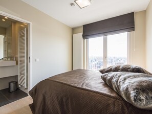 A bedroom with a bed, a window with a view, a radiator, and a door leading to a bathroom. - Beautiful apartment by the sea (Blankenberge)