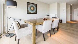 A modern dining area with a wooden table, white upholstered chairs, and a large abstract painting on the wall. - Beautiful apartment by the sea (Blankenberge)