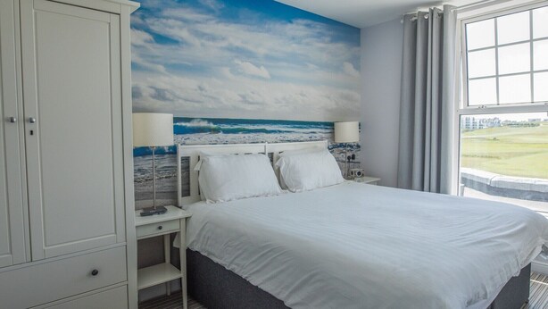 Une chambre d’hôtel comprenant un lit, une table de chevet, une armoire et une grande fenêtre donnant sur la mer et un champ verdoyant.