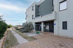 A modern building with a flat roof, a swing set, and a paved walkway. - Apartment Sang Near The Beach (Pula)