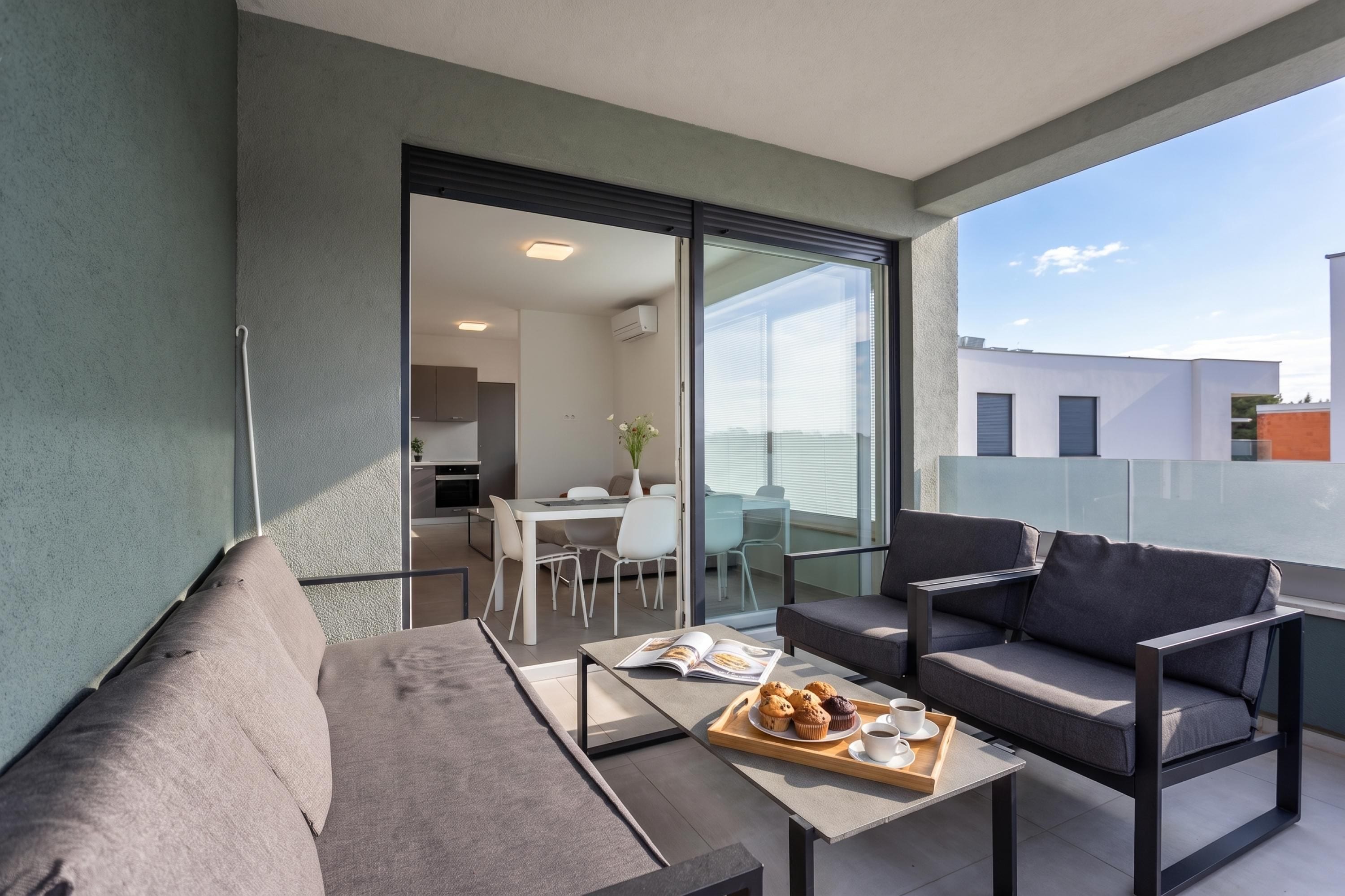 A modern balcony with a grey sofa, a small table with a tray of muffins and a cup, and a glass door leading to a dining area.