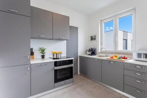 A modern kitchen with grey cabinets, a built-in oven, a coffee machine, and a window with a view of the sky. - Apartment Sang Near The Beach (Pula)
