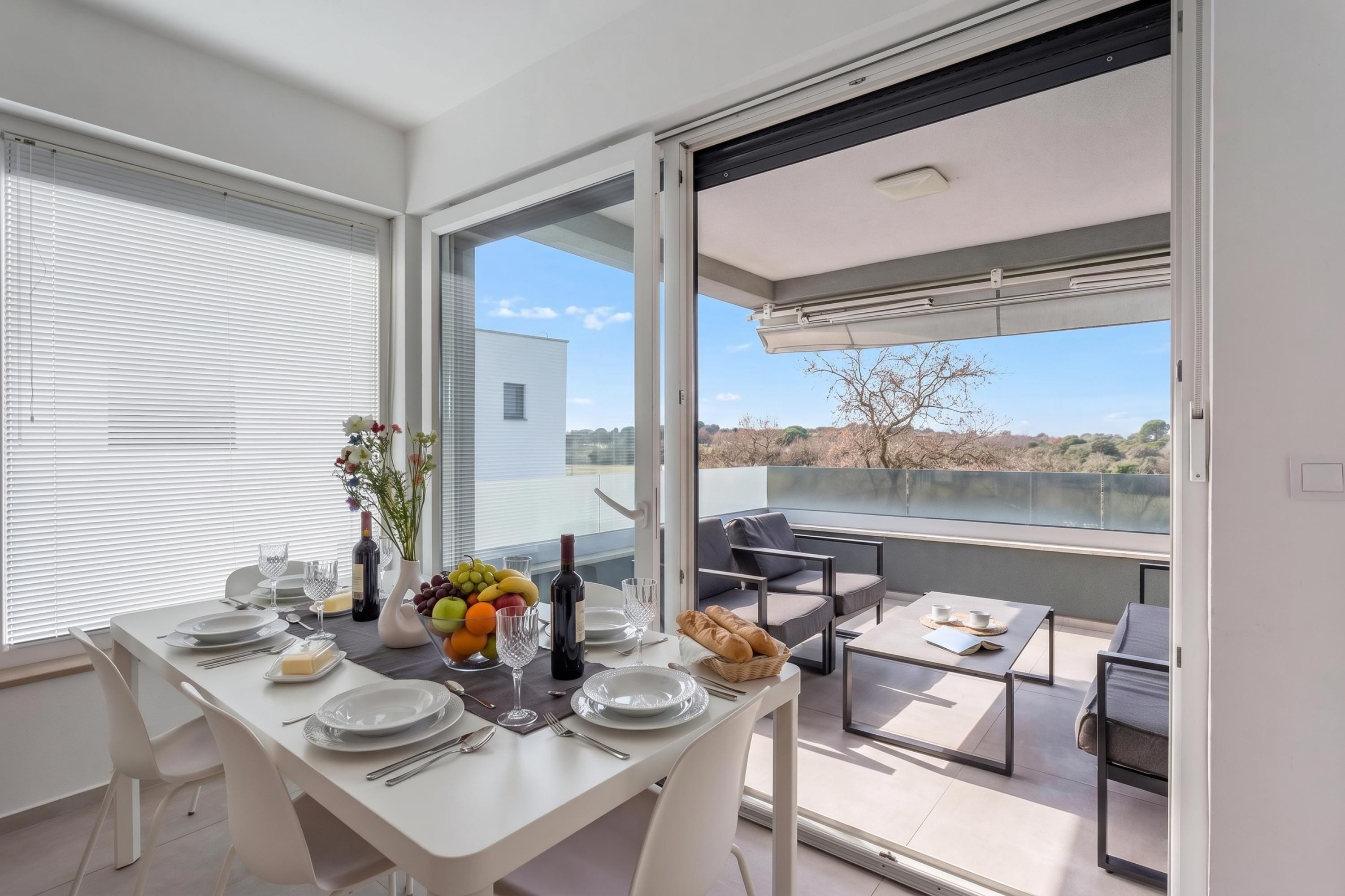 Uma sala de jantar com mesa posta para quatro pessoas, vista para o exterior e uma varanda com assentos.