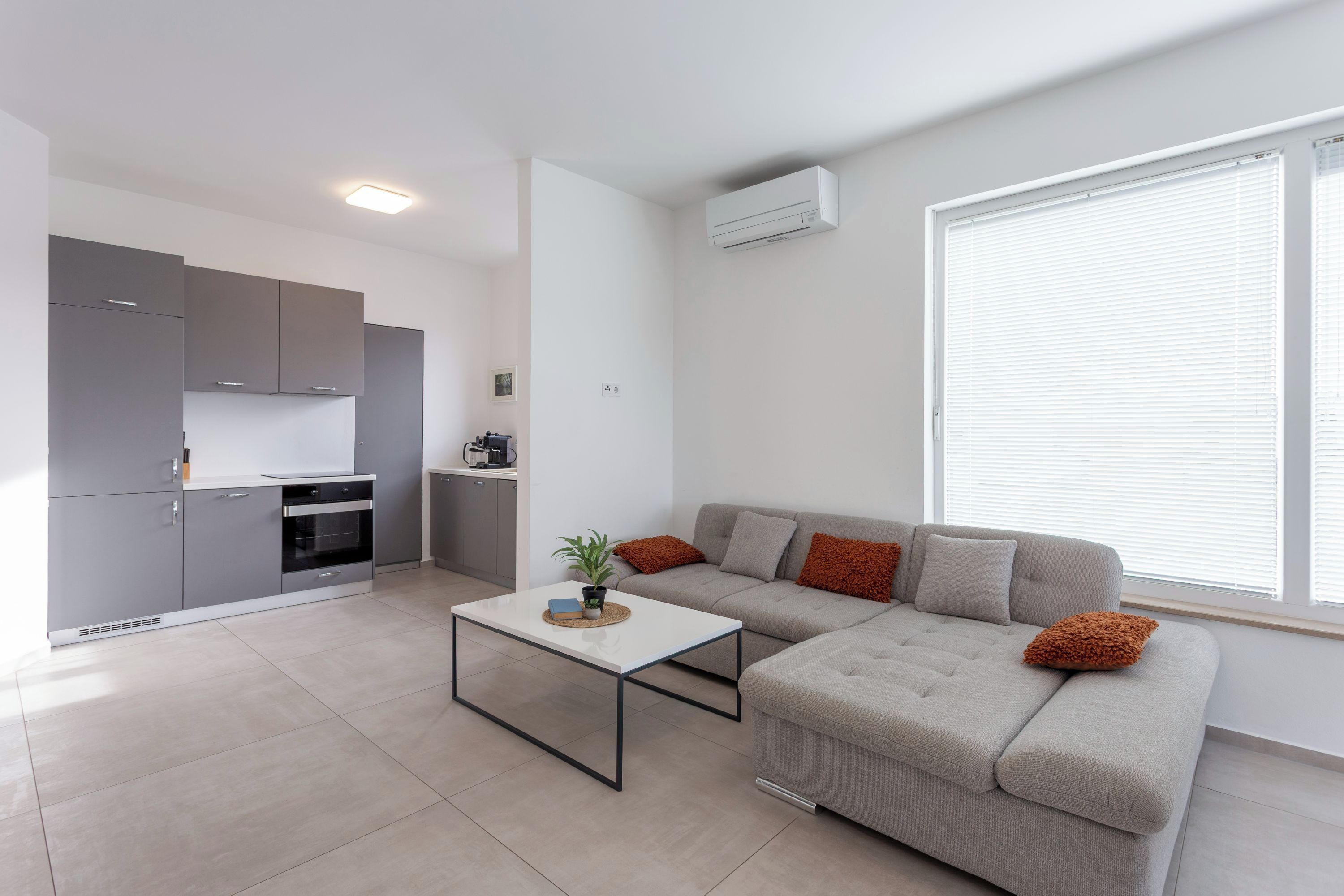 Un espace de vie moderne comprenant une cuisine, un canapé gris agrémenté de coussins, une table basse et une grande fenêtre avec des stores.