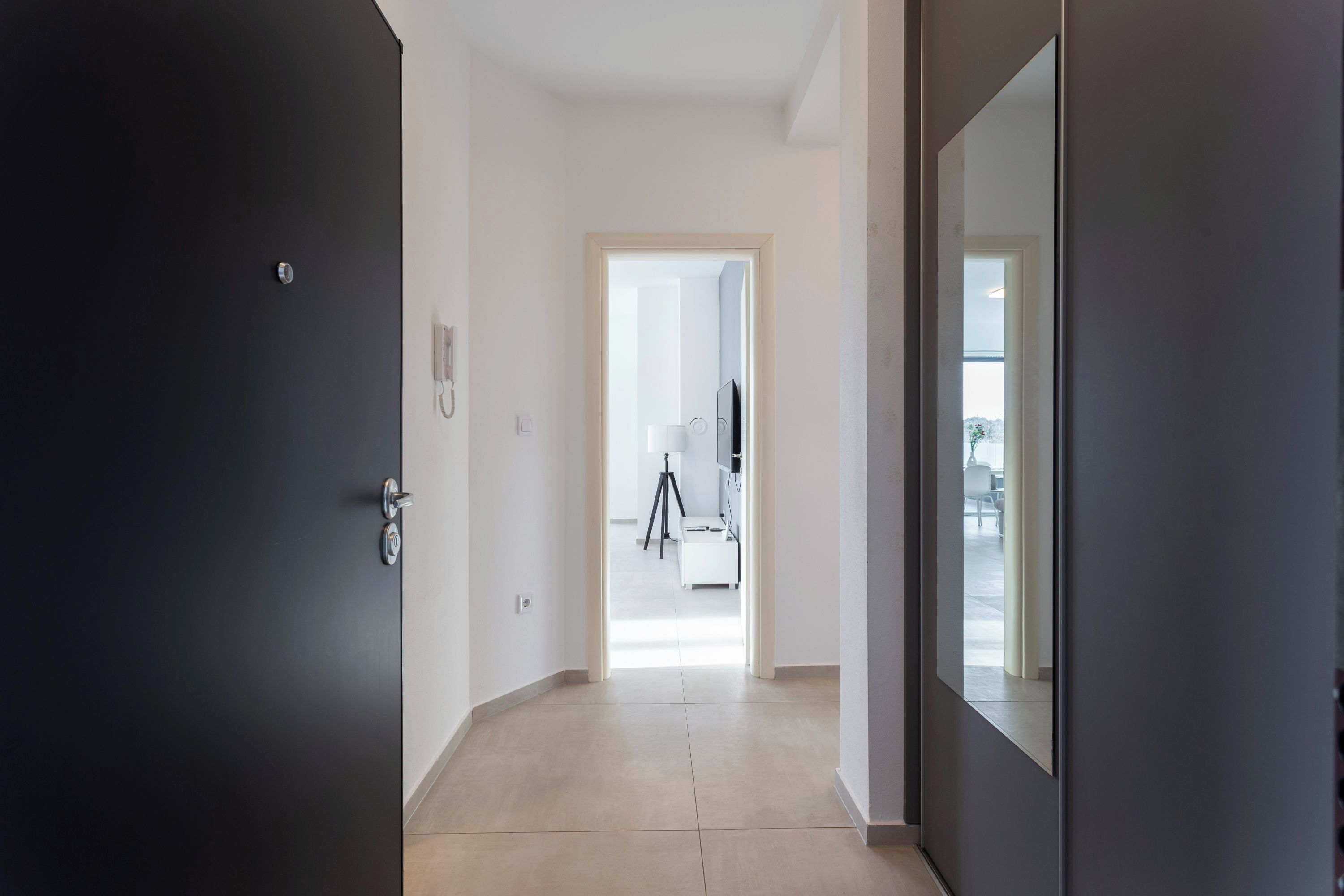 A hallway with a dark door on the left, a light-colored floor, and a mirror on the right. A door at the end of the hallway leads to a room with a tripod and a white chair.