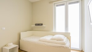 Um quarto pequeno e bem organizado, com uma cama de solteiro, uma mesinha de cabeceira, um armário pequeno e uma janela com persianas.
