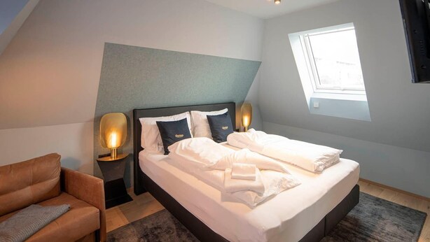 A modern bedroom with a large bed, two bedside tables with lamps, a sofa, and a window providing natural light. - Backhome City Apartments (Innsbruck)