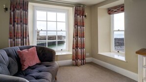 A room with a grey sofa, two windows with curtains, a view of the sea, and a bench by the window. - Rock House Hotel - Guest House (Lynmouth)