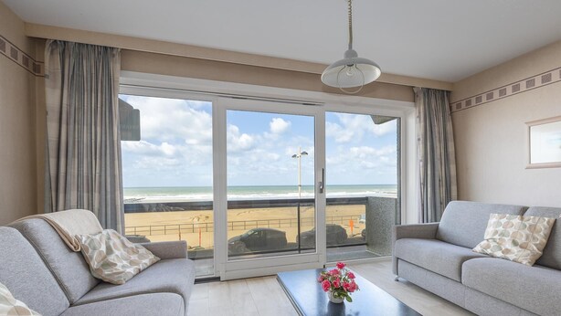Sala de estar com sofá, mesa de centro com flores e vista para a praia através de grandes portas de vidro.