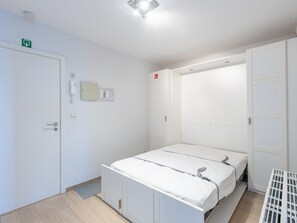 Une pièce propre et moderne, avec un lit, une armoire blanche, une porte portant un panneau indiquant la sortie de secours, et un petit panneau de commande au mur.