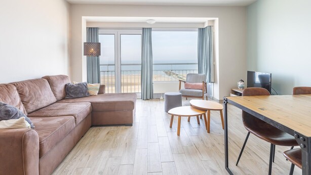 Una sala moderna con un sofá de cuero marrón, una mesa de comedor de madera con sillas y un gran ventanal con vista a la playa.