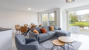 Un salon moderne avec un canapé d’angle gris, deux fauteuils, une table à manger avec des chaises et une grande fenêtre donnant sur l’extérieur.