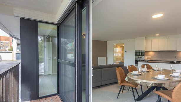 Un appartement moderne doté d’un balcon, d’un coin repas avec une table ronde et des chaises, et d’une cuisine avec des meubles blancs.