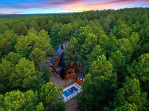 A secluded cabin surrounded by dense forest, with a swimming pool and outdoor seating area. - Designer Cabin | Heated Pool, Hot Tub & Firepit (Broken Bow)