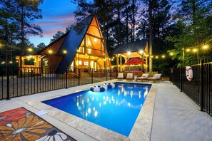 A pool area with a wooden deck, lounge chairs, and a building with a sloped roof.