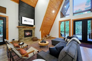 Un salon spacieux avec une cheminée, un grand canapé et une vue sur la forêt par les fenêtres.