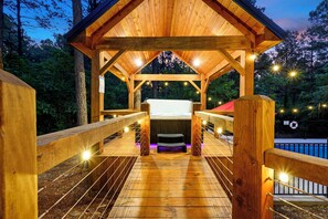 A wooden deck with a covered seating area, a bench, and a pool in the background. - Designer Cabin | Heated Pool, Hot Tub & Firepit (Broken Bow)