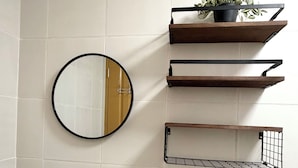 Een badkamer met een wit wastafel, toilet en een ronde spiegel. Daarboven, boven het toilet, zijn houten planken en op de bovenste plank staat een potplant.