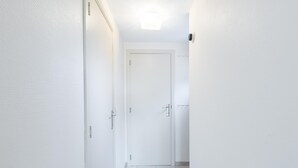 Kapea käytävä, jonka seinät ovat valkoiset ja jonka lattia on laatoitettu, johtaa suljettuun oveen.