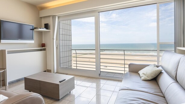 Ein modernes Wohnzimmer mit einem großen Fernseher, einer Couch, einem Couchtisch und Blick auf den Strand durch eine Schiebetür.