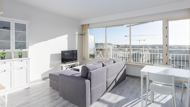 Un salon moderne avec un canapé gris, un téléviseur et un coin repas comprenant une table et des chaises blanches. De grandes fenêtres offrent une vue sur le paysage urbain.