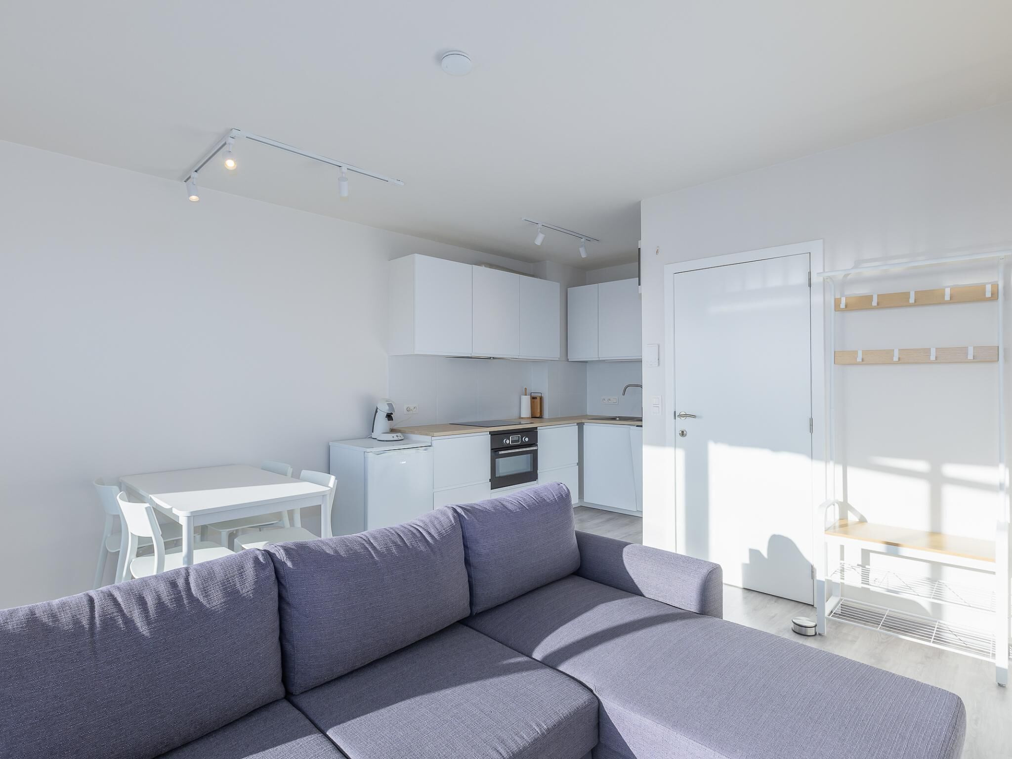 Un appartement moderne avec un canapé gris, une table à manger blanche et un coin cuisine doté de meubles blancs et d’un plan de travail en bois.