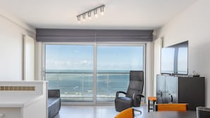 A modern hotel room with a large sliding glass door to the sea, a dining area with a round table, and a flat-screen TV mounted on the wall. - Charming studio on the seafront with sea view (Middelkerke)