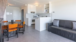 A modern kitchen with white cabinets, a microwave, and a kettle. There is a dining table with orange chairs and a grey sofa. - Charming studio on the seafront with sea view (Middelkerke)