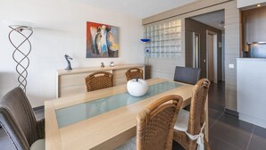Een moderne eethoek met een tafel met glazen blad, rieten stoelen, een schilderij aan de muur en een keuken op de achtergrond.