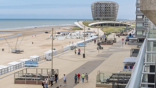 Een boulevard aan zee met een opvallend bolvormig gebouw, een zandstrand en een promenade met zitjes en een voetpad.