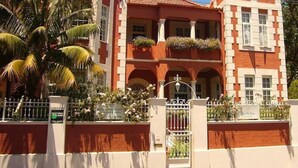 Un immeuble de deux étages avec une façade rouge et blanche, des balcons ornés de jardinières et un palmier au premier plan.