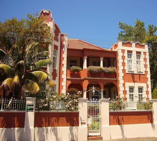 A two-story building with a red and white facade, balconies with flower boxes, and a palm tree in the foreground. - The Villa Rosa Bed and Breakfast (Cape Town)