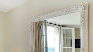 Habitación de hotel con un espejo grande y ornamentado, un cabecero de madera y vista al mar a través de una ventana abierta.