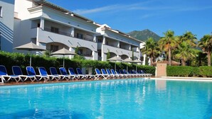 A swimming pool with blue lounge chairs and umbrellas, a white building with balconies, and palm trees in the background.
