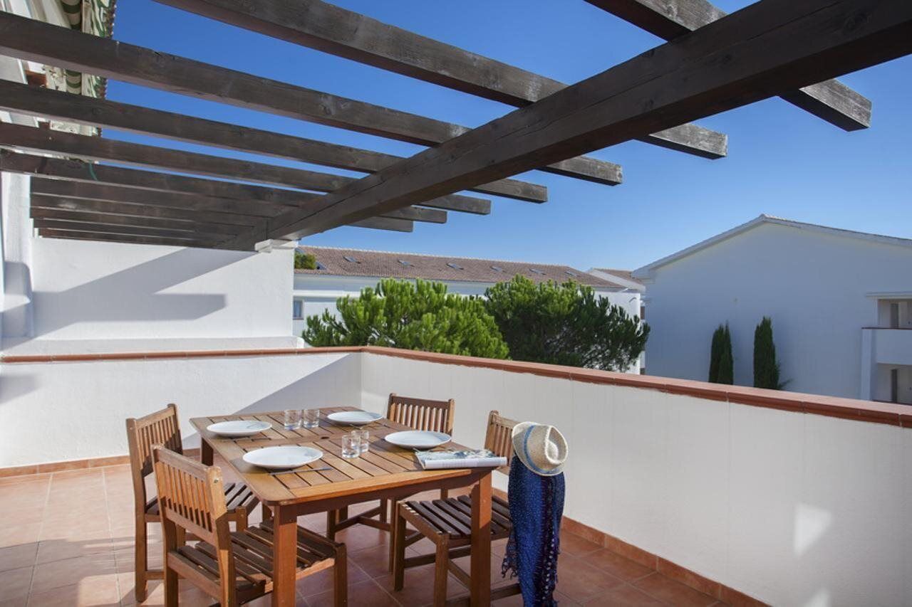 A wooden table set for four with plates and glasses, a hat, and a view of a building and greenery in the background.