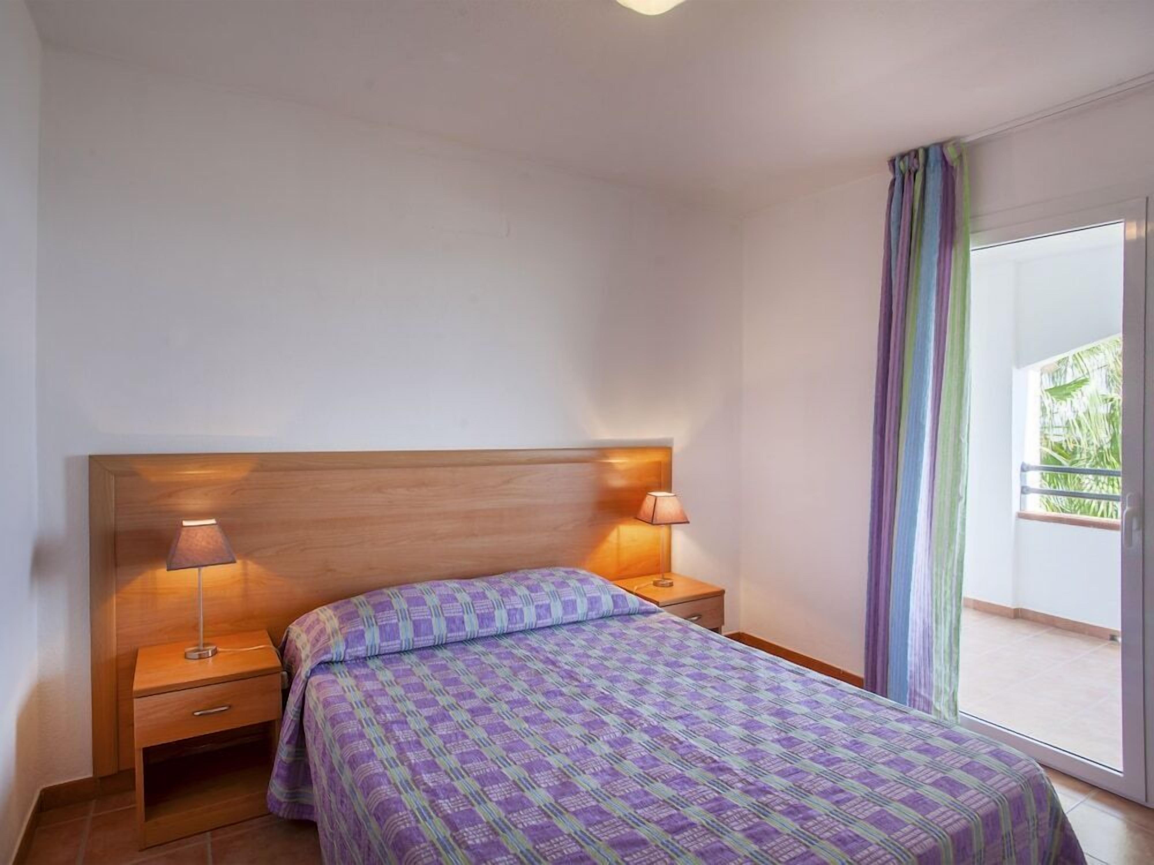 A bedroom with a wooden headboard, two bedside lamps, a patterned bedspread, a nightstand with a drawer, and a view through a window with curtains.