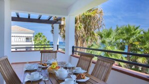 A wooden table set for breakfast with white plates, cups, and a fruit bowl, overlooking a beach with palm trees and a building in the background.