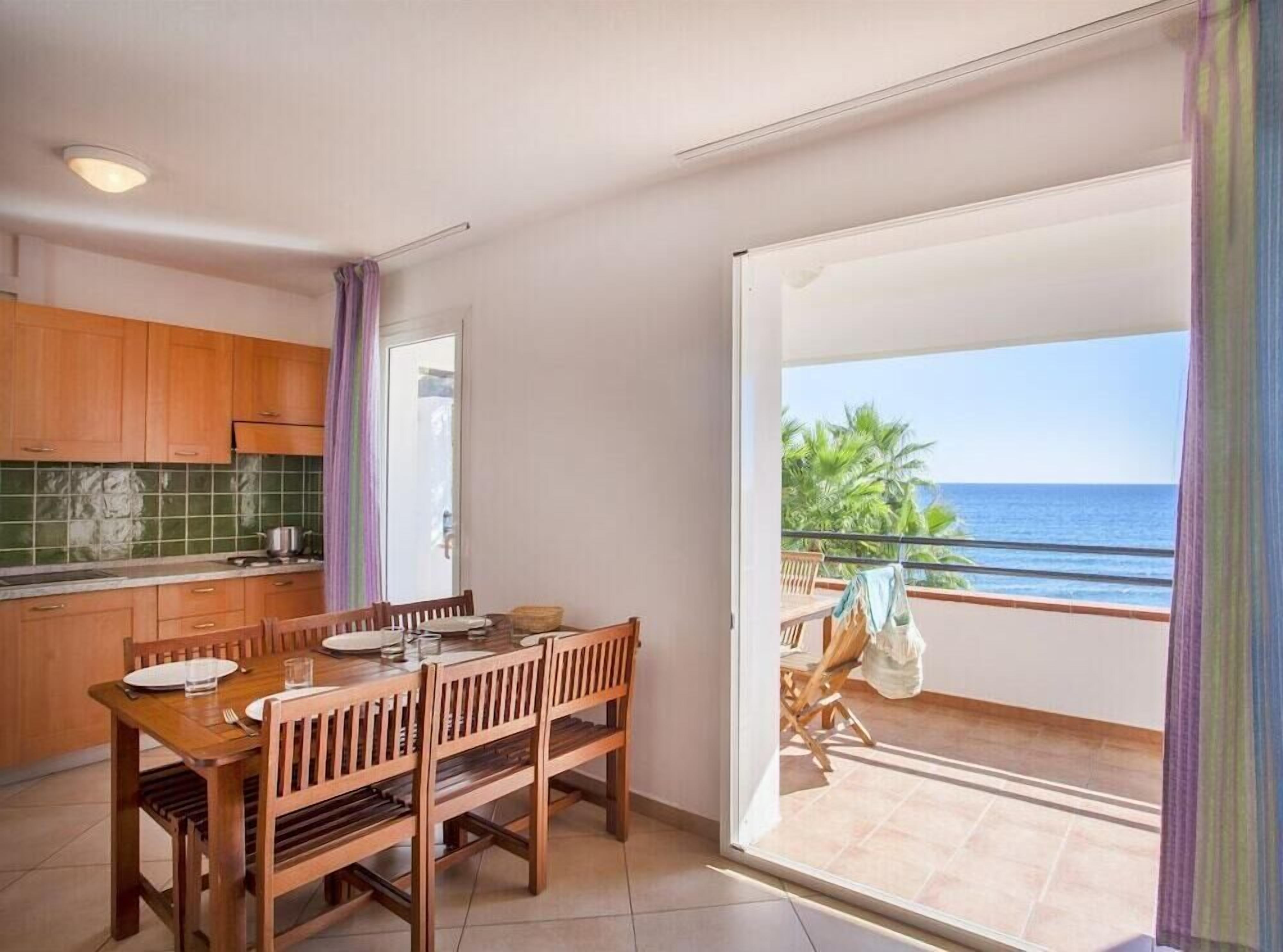 A kitchen with wooden cabinets, a dining table with chairs, and a view of the ocean through a sliding glass door.