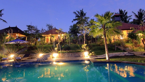 A pool area with a clear view of the pool, surrounding palm trees, and a building with a red roof. - Villa Capung Mas (Ubud)