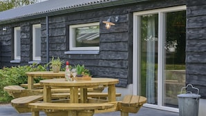 Ein hölzerner Essbereich im Freien mit Tisch und Bänken, einem Mülleimer und einem Haus mit dunkler Fassade und großen Fenstern.