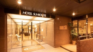 La entrada del Hotel Sunroute, con fachada de vidrio, un mostrador de recepción y una planta en maceta en el exterior.