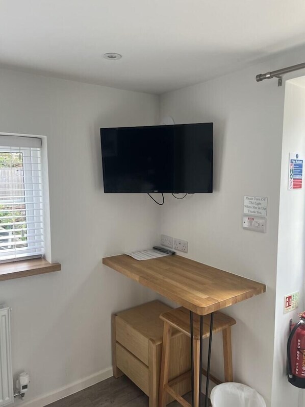 A small room with a wooden desk, a mounted TV, a window with blinds, and a fire extinguisher on the wall. - The Priory Burford Bed and Breakfast (Burford)