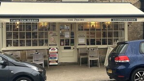 Un café con un letrero que dice 'The Priory' y 'Café Tea Rooms'. Hay áreas de estar al aire libre con sillas y mesas.