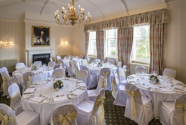 A formal dining room with round tables set for a meal, white chairs with gold sashes, a chandelier, and a fireplace. - The Talbot Hotel (Malton)