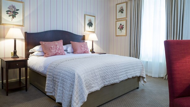 Une chambre d’hôtel avec un grand lit, deux tables de chevet avec des lampes, un fauteuil rouge et des œuvres d’art florales accrochées au mur.