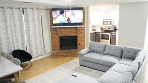 Una sala de estar con un sofá gris, un televisor montado sobre una chimenea de ladrillo, una mesa de comedor con sillas y una cocina al fondo.