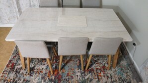 Una mesa rectangular de mármol con un camino de mesa tejido, rodeada por seis sillas tapizadas de color gris con patas de madera.