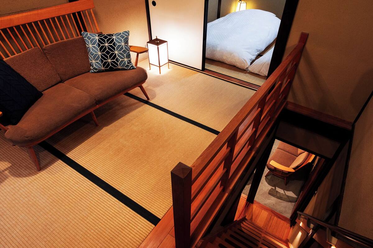 Ein traditioneller japanischer Raum mit Tatami-Böden, einem niedrigen Holzbett, einer Couch mit Kissen und einem kleinen Tisch mit Lampe.