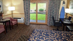Ett hotellrum med en säng, ett skrivbord, en stol, ett litet bord, en lampa och utsikt över en trädgård genom en skjutdörr.